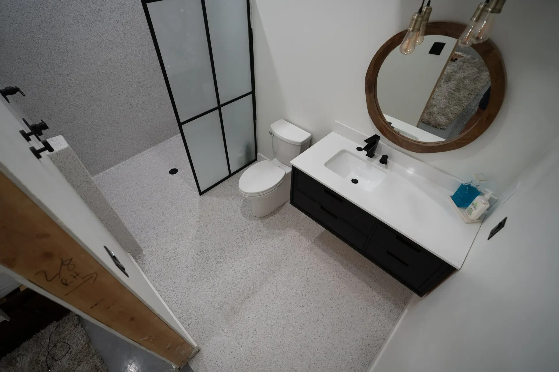 Modern bathroom with white speckled floor, black vanity, glass shower, and round mirror.