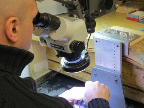 Person using a microscope to inspect a metal part on a workbench under bright light
