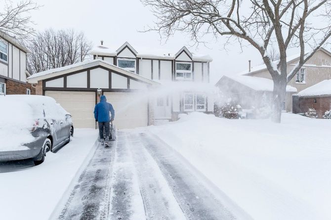 Snowy suburban driveway being cleared by a person in a blue jacket, with snow-covered houses and cars.