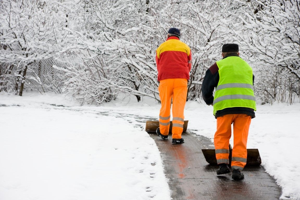 Two road workers in orange uniforms clearing a snowy path through a winter forest
