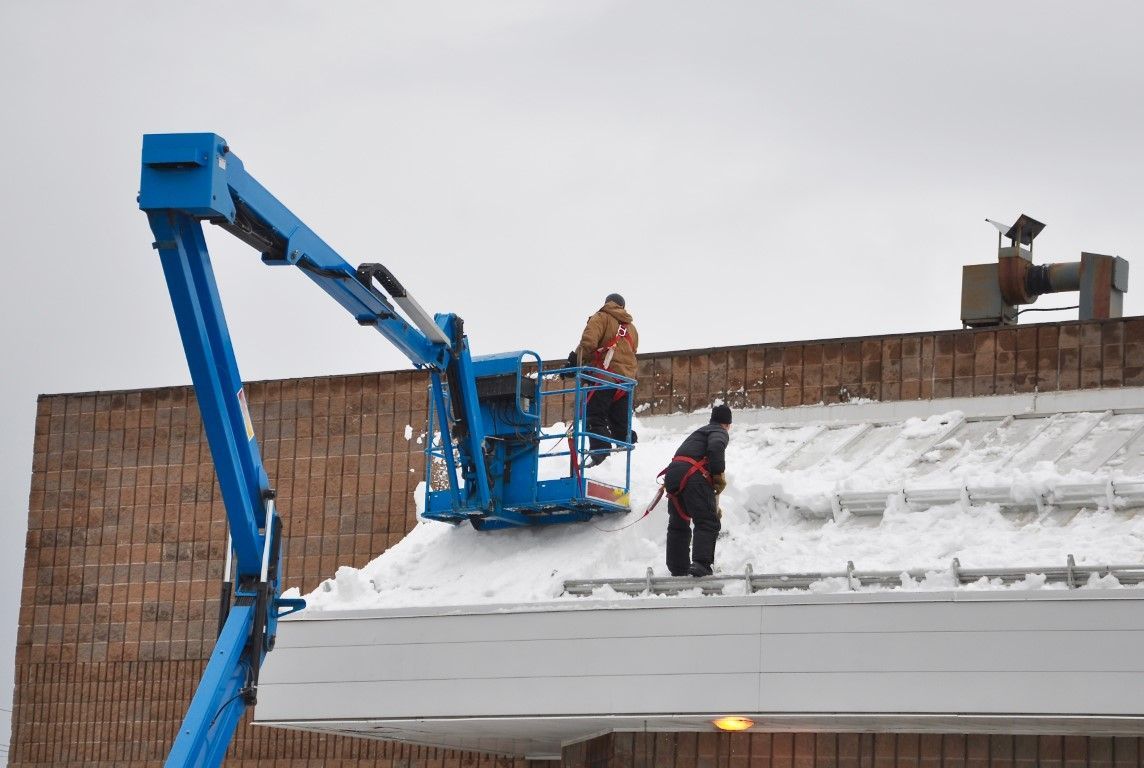 Workers in a blue boom lift clear snow from a rooftop beside a brick wall.