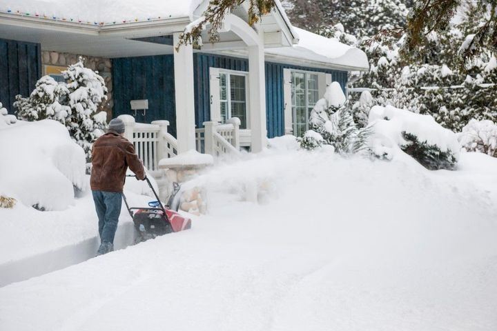 Person using a snow blower in a snow-covered yard beside a blue house