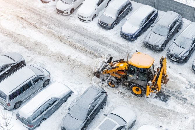 Snowplow clearing a snowy parking lot beside rows of parked cars