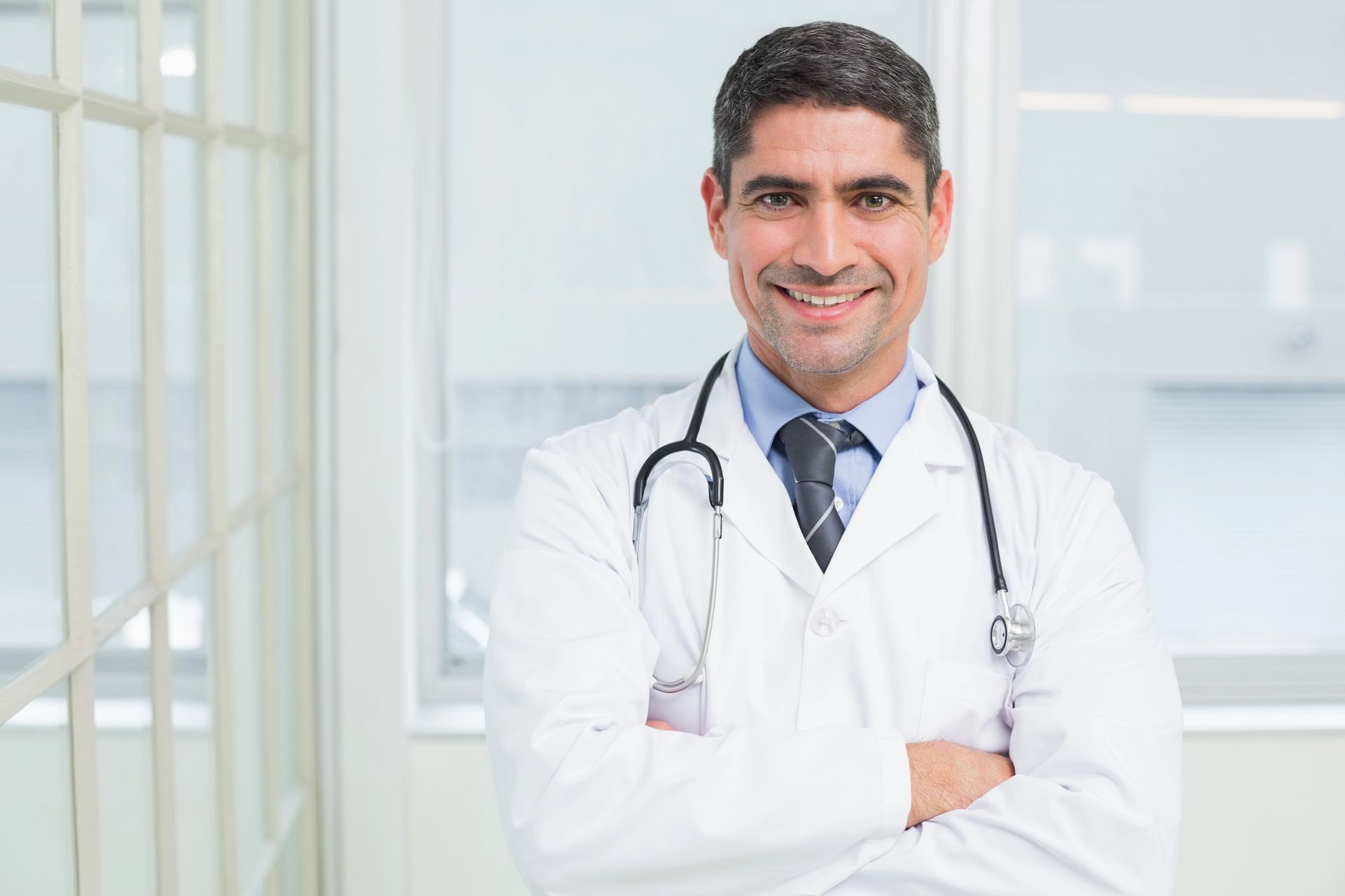 Médico con bata blanca y estetoscopio de pie frente a un edificio, con los brazos cruzados y sonriendo.