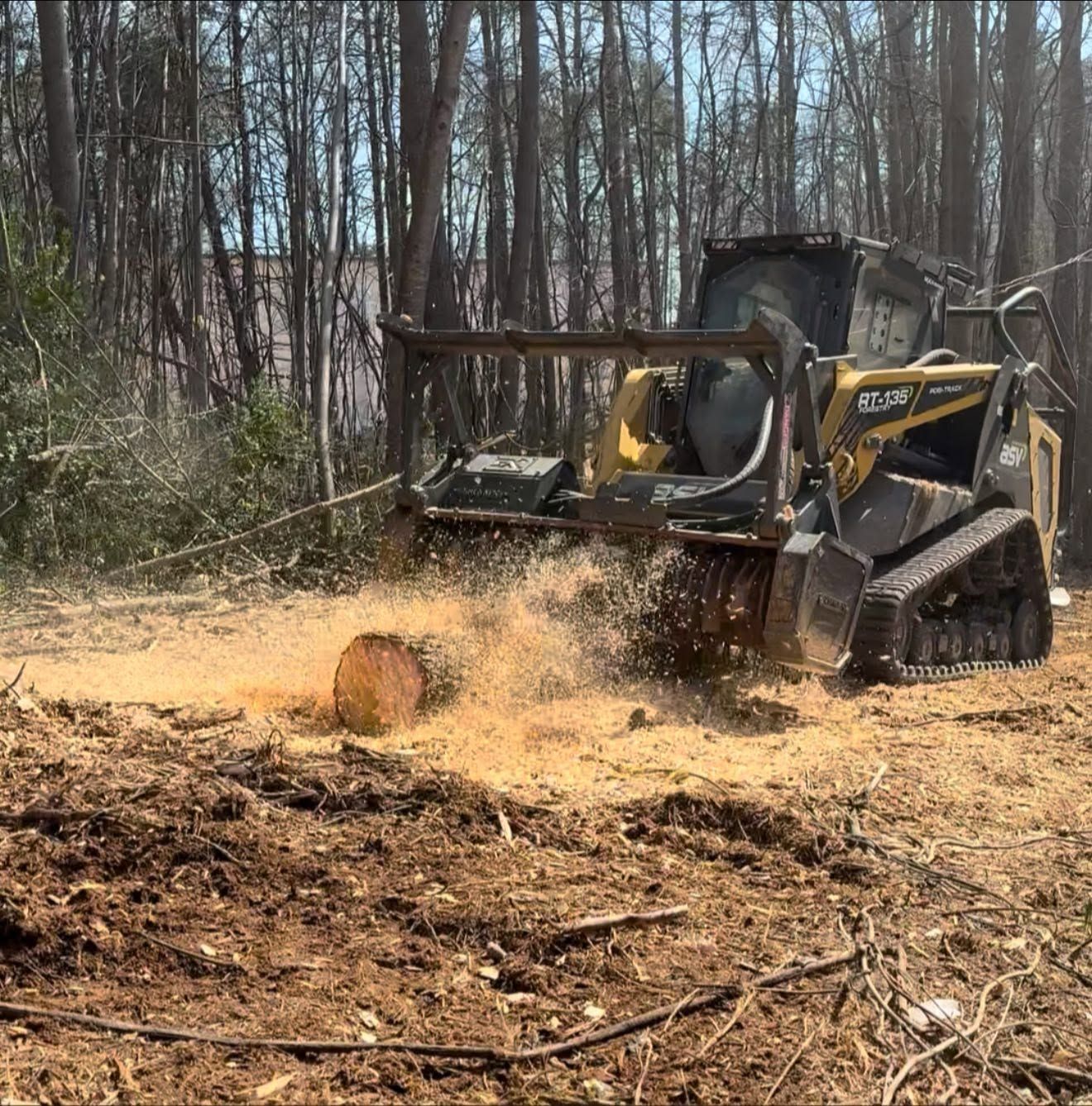 Skid steer clearing brush in a wooded area, kicking up wood chips and debris