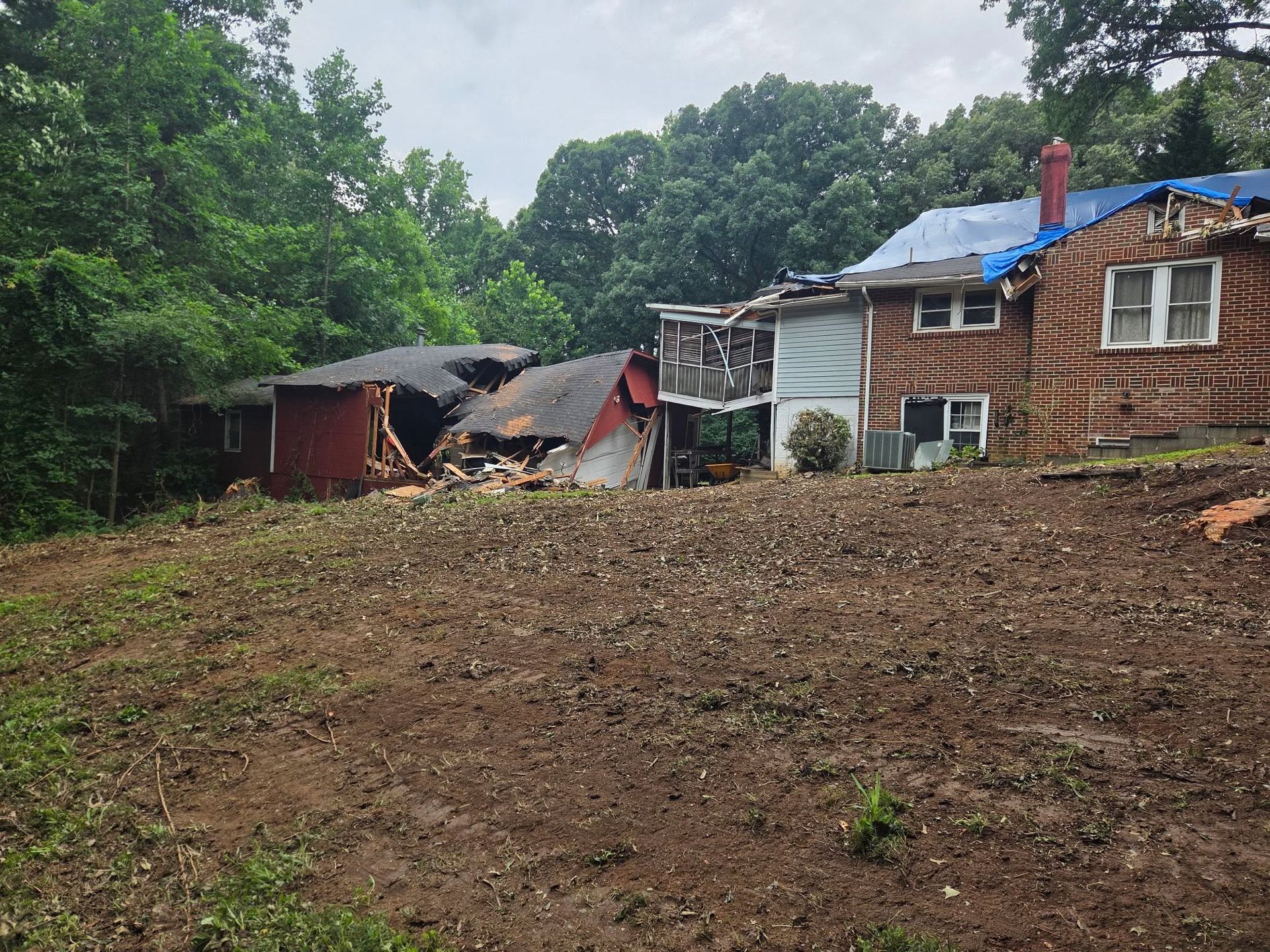 Collapsed rural house beside a muddy slope and trees, with a brick home and blue tarp roof nearby.