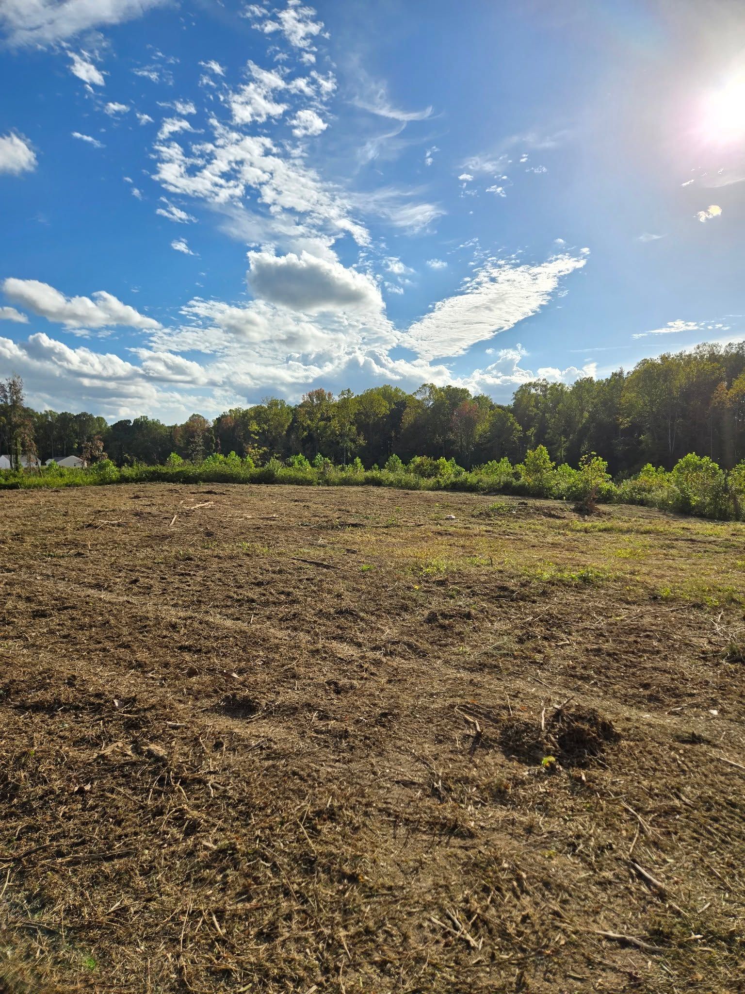 Sunny plowed field under a blue sky with scattered clouds and a tree line in the distance