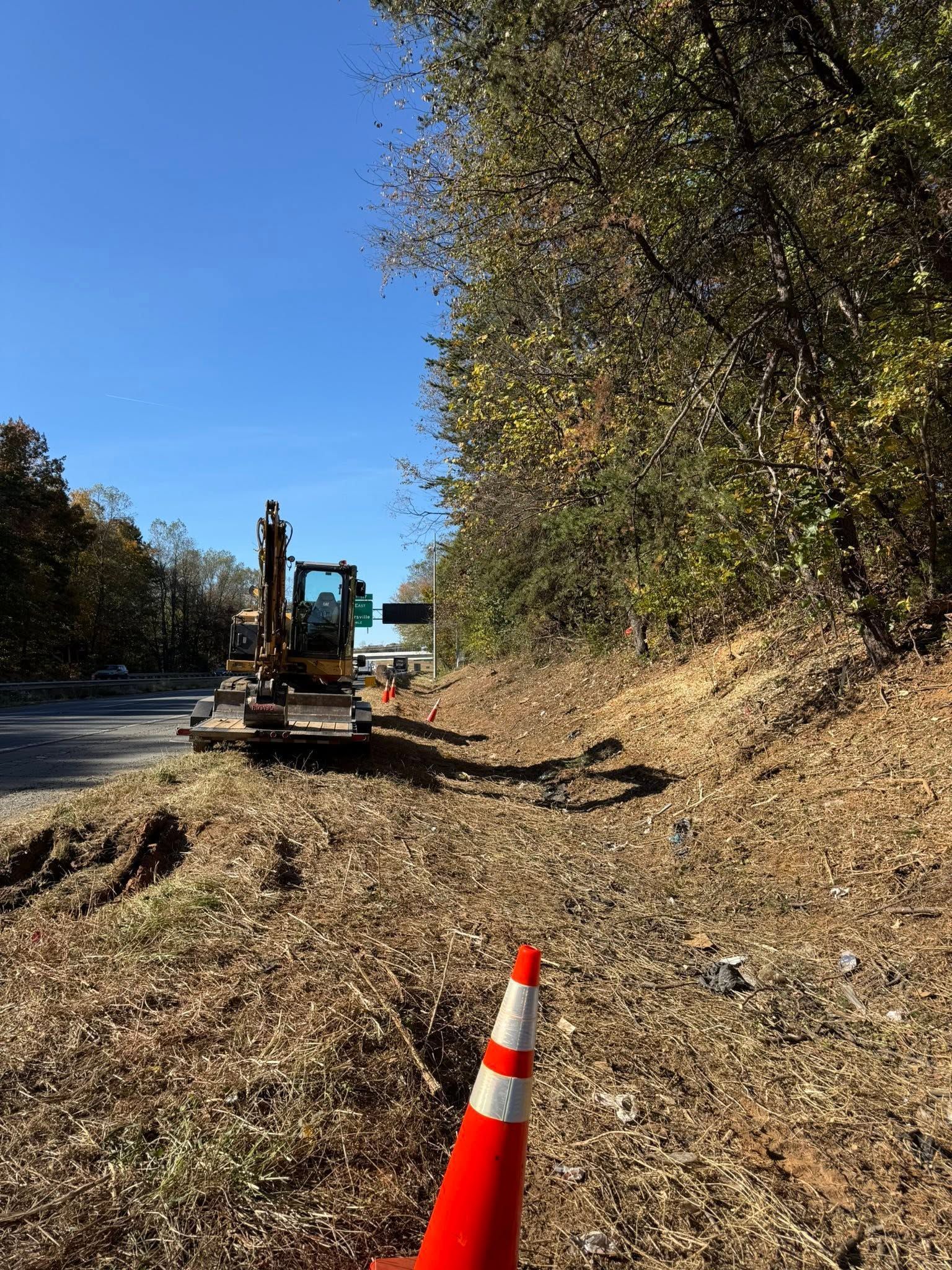 Excavator on a leaf-covered roadside with orange cone, beside a steep wooded slope under a blue sky