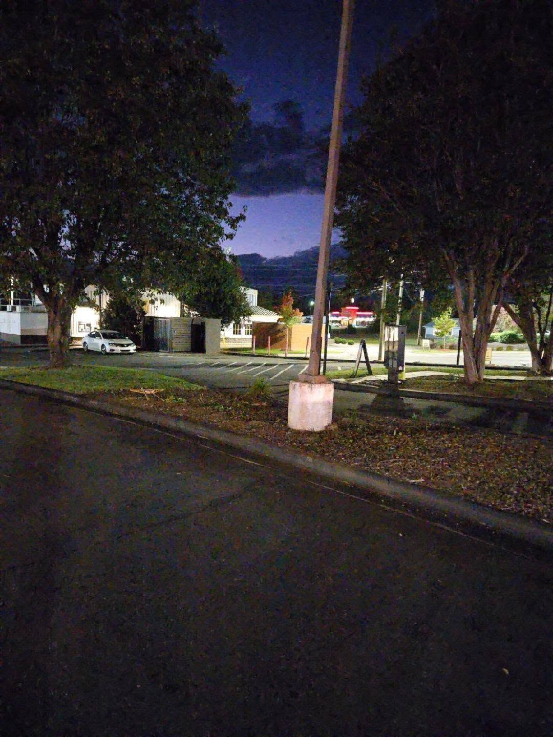 Dusk-lit parking lot with trees, buildings, and a road sign in the center.