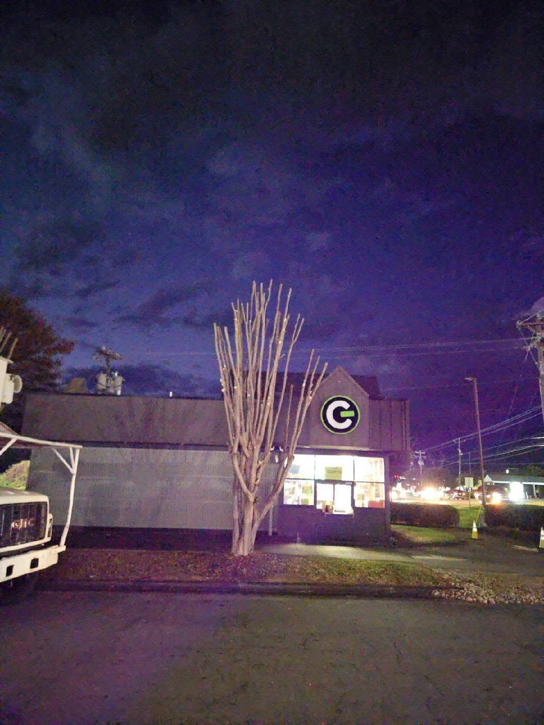 Night view of a building with a circular logo, lit windows, parked truck, and a bare tree under a purple sky