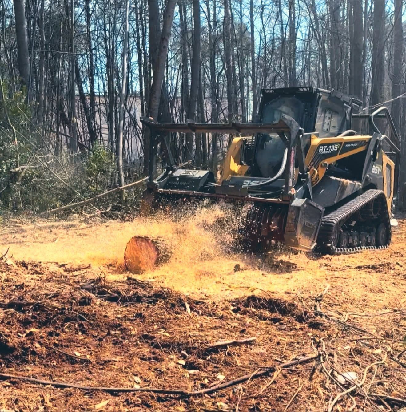 Yellow skid-steer mulching a stump in a wooded clearing, kicking up wood chips.