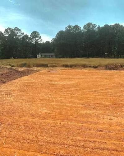Freshly graded orange dirt lot with a grassy field and tall trees under a blue sky.