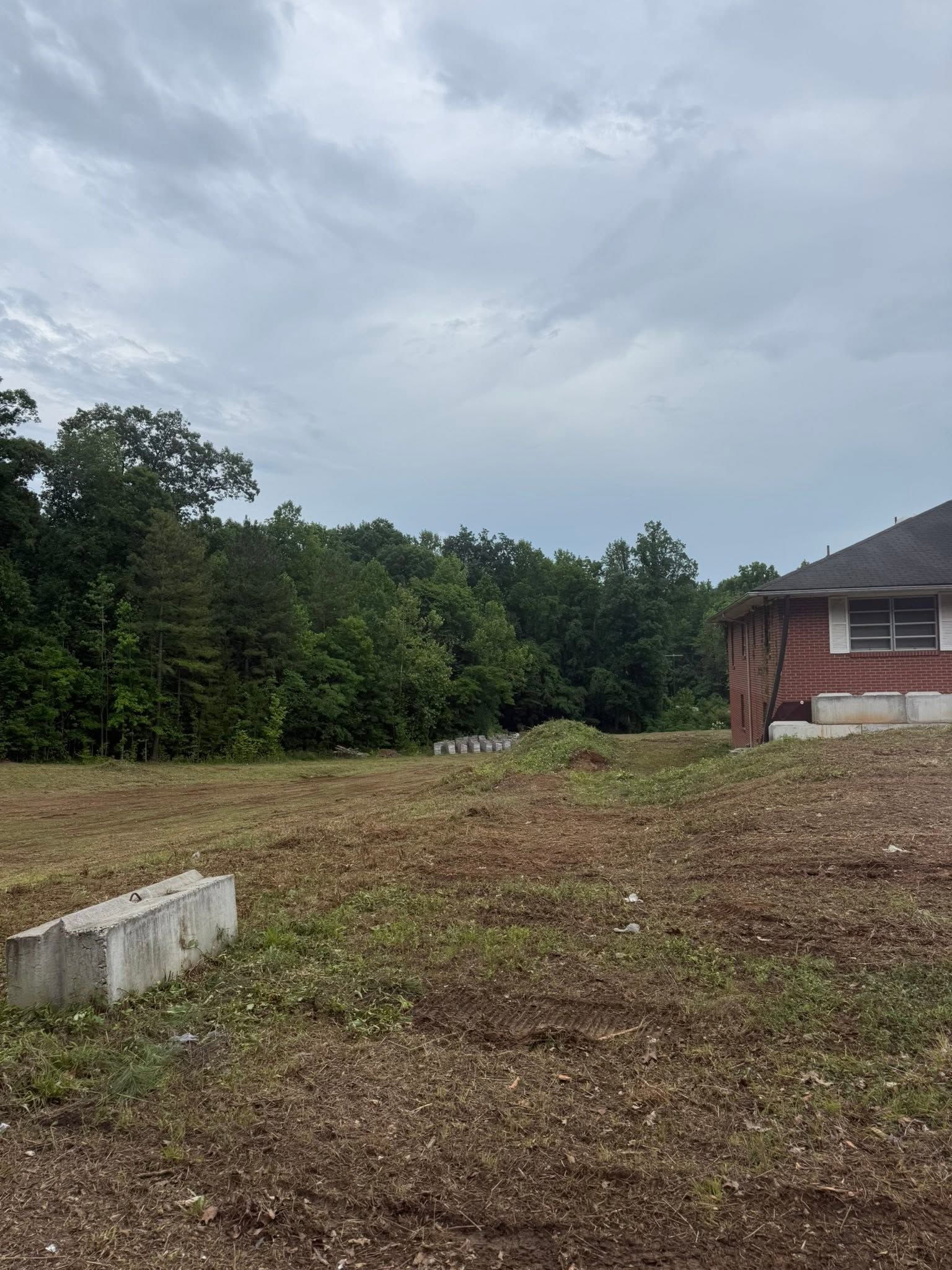 Empty dirt lot beside a brick house, with trees under a cloudy sky