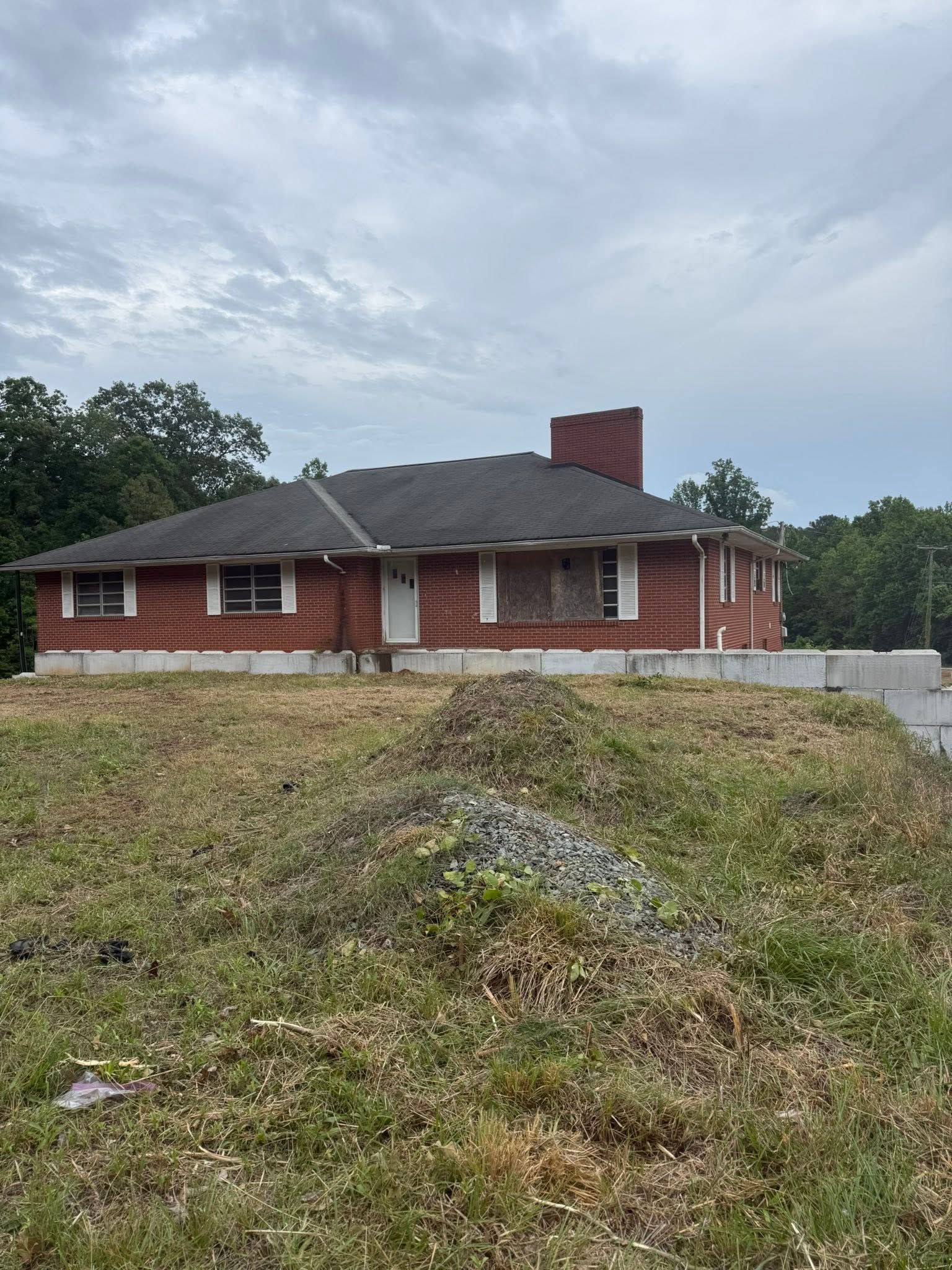 Single-story red brick house with a dark roof on a grassy, overgrown lot under a cloudy sky