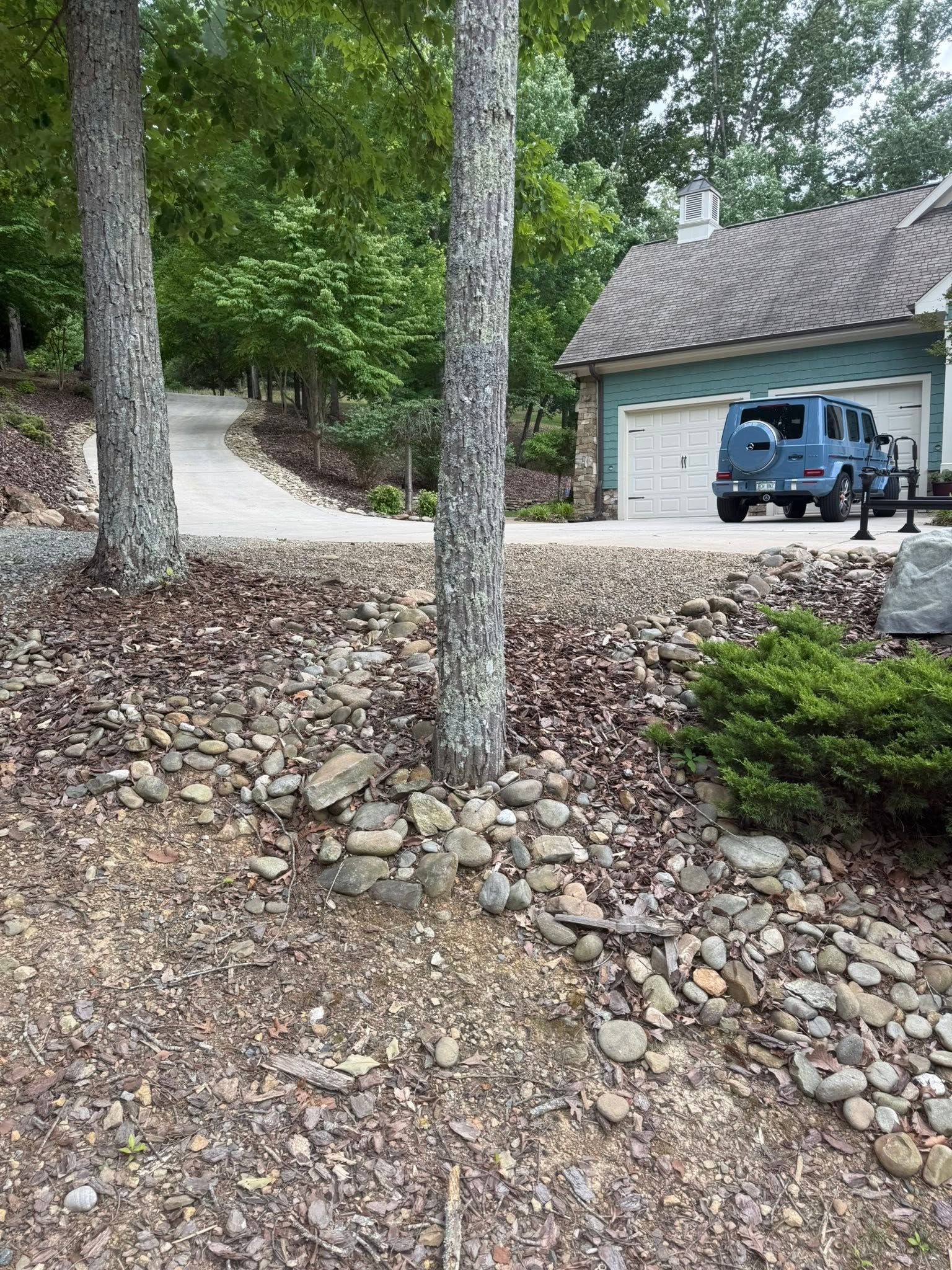 Rocky wooded yard with trees, driveway, and blue SUV parked beside a garage