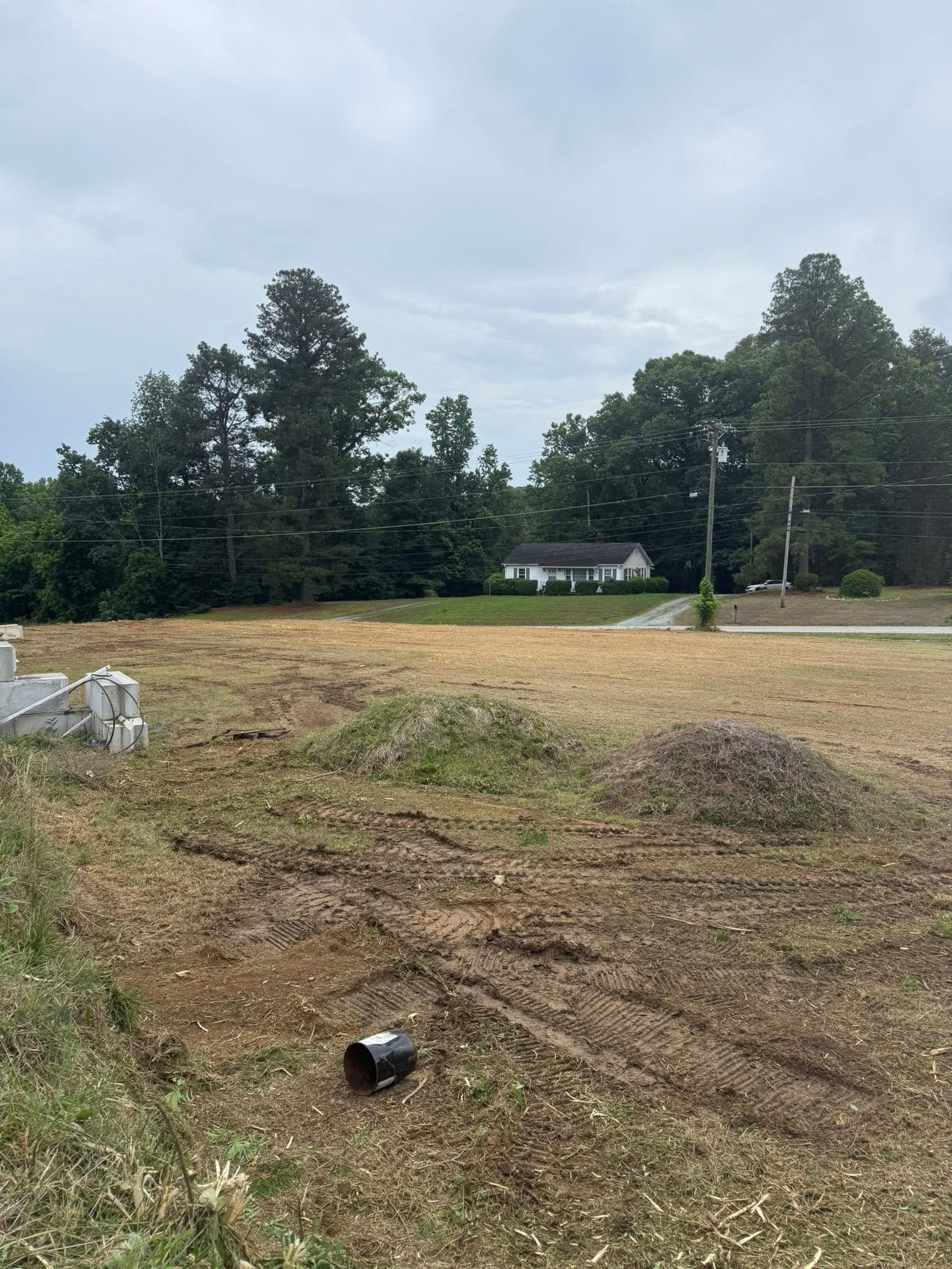 Cleared dirt lot with trees and a house in the background under a cloudy sky