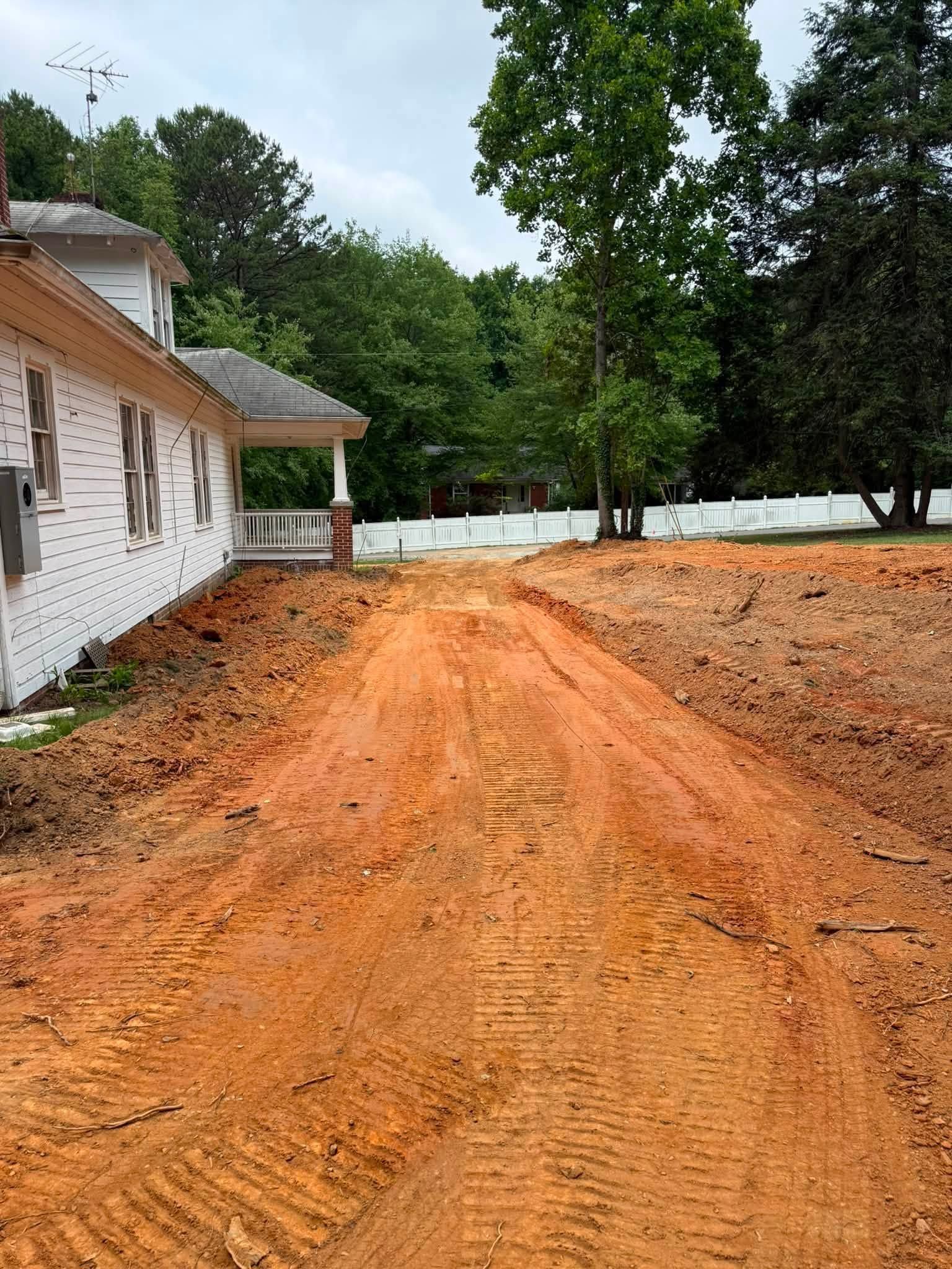 Red dirt driveway beside a white house, leading through a wooded yard on an overcast day