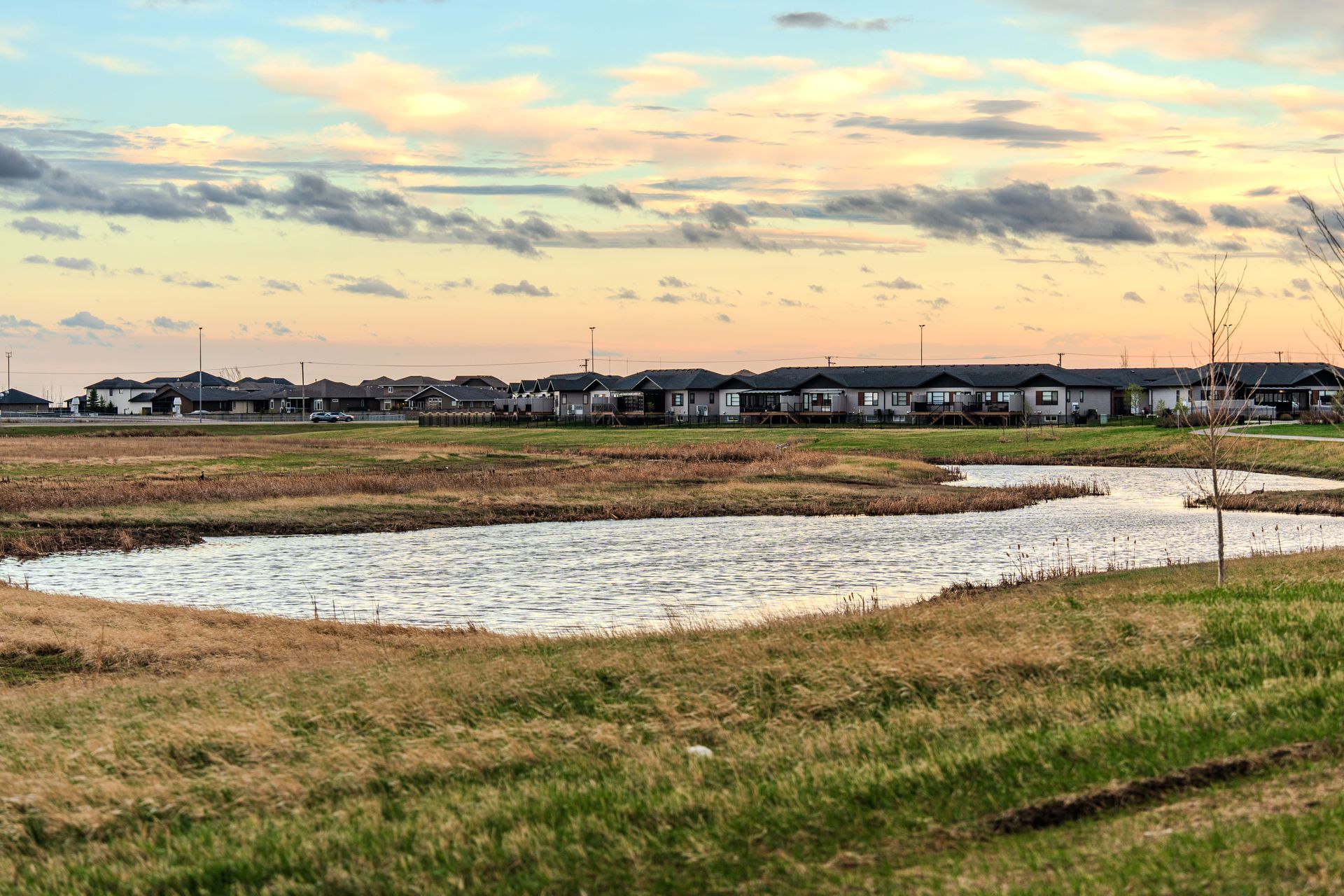 Grassy field with a shallow pond at sunset, with houses along the horizon under a cloudy sky