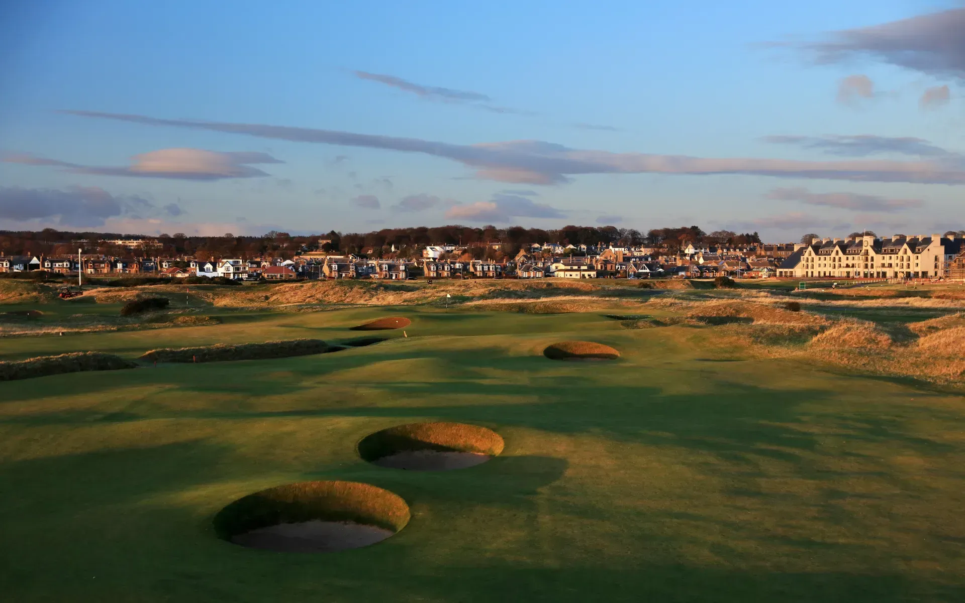Sunlit golf course with sand bunkers, green fairway, and coastal town under a partly cloudy sky