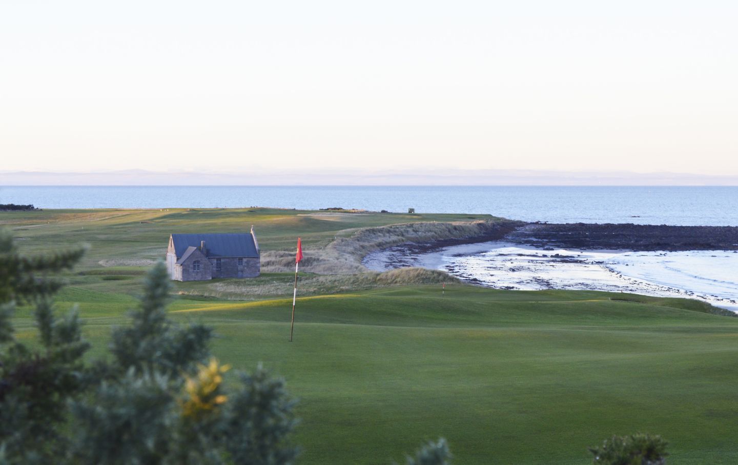 Golf course by the sea with a small building and red flag at sunset