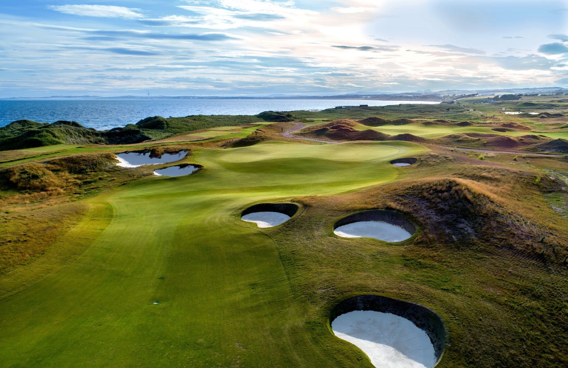 Scenic coastal golf course with green fairway, sand bunkers, and ocean under a cloudy sky