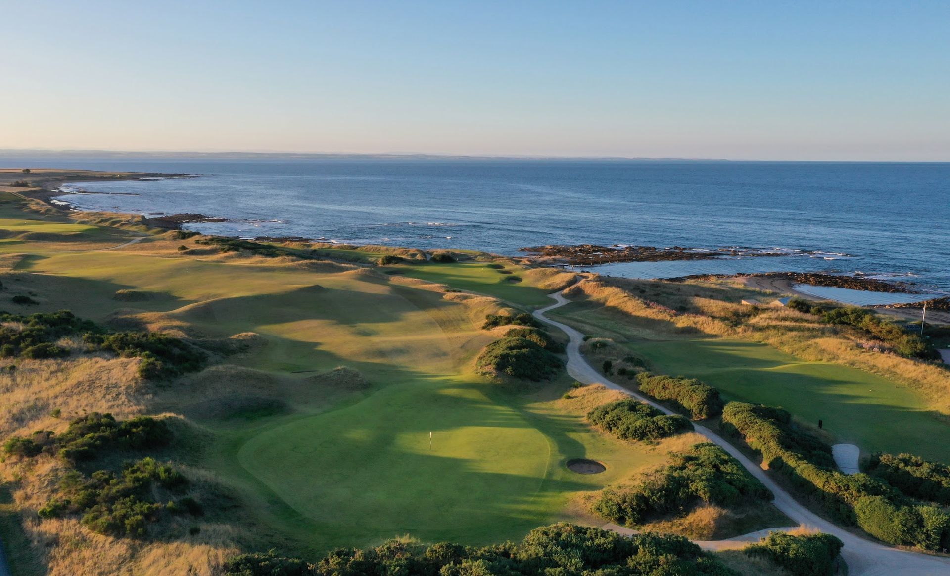 Coastal golf course with green fairways and sandy dunes beside the blue ocean at sunset