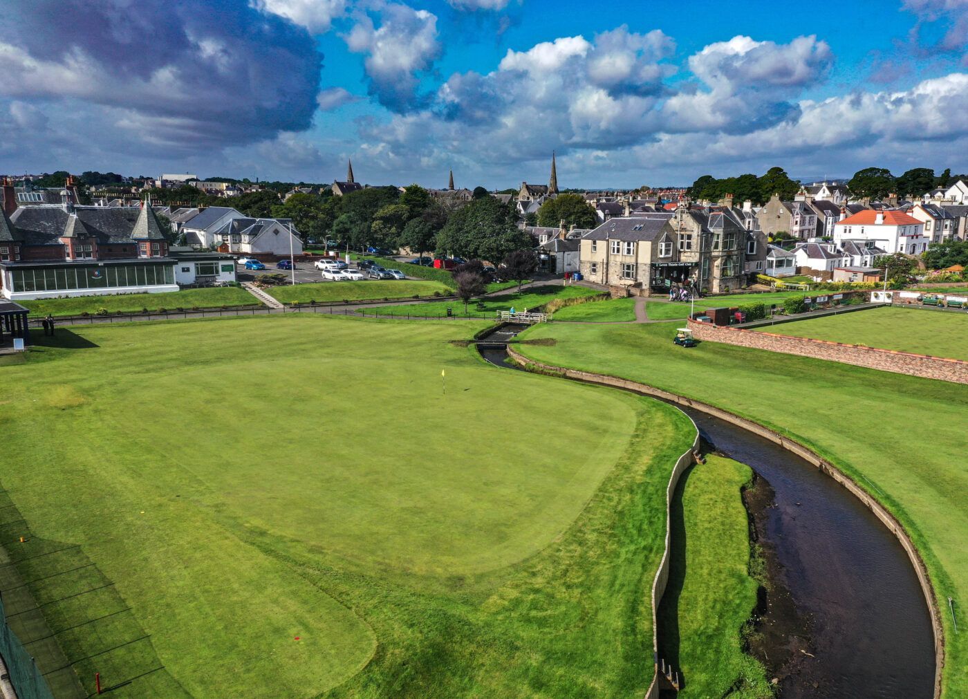 Green golf course beside a winding canal, with houses and cloudy sky in the background