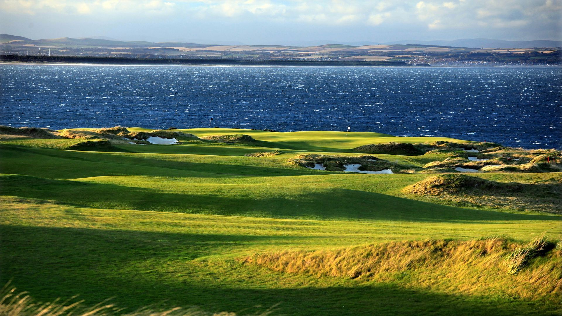 Green coastal golf course beside a deep blue bay under a bright, windy sky