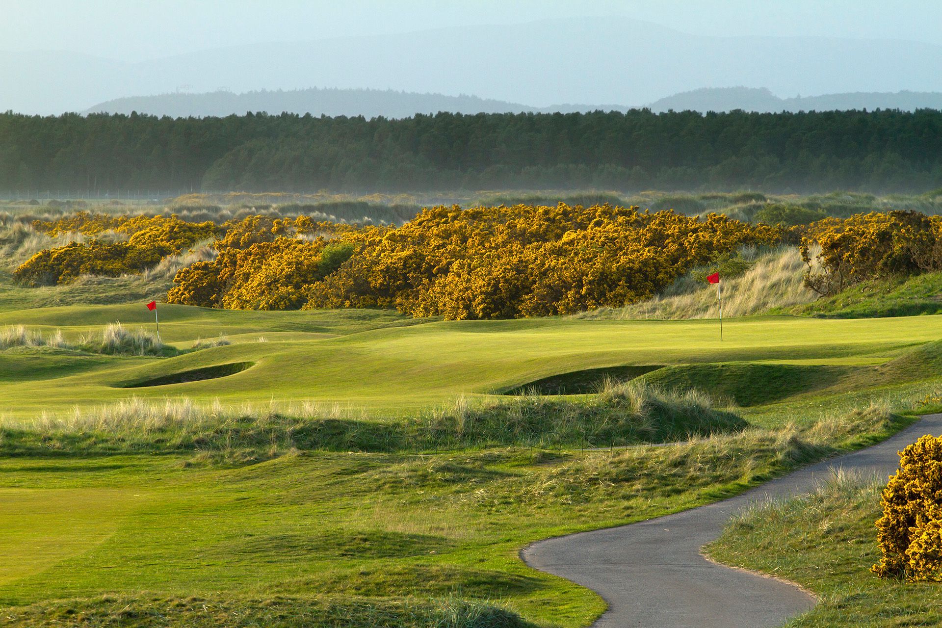 Golf course with green fairways, sand bunkers, yellow shrubs, and a winding path under a hazy sky