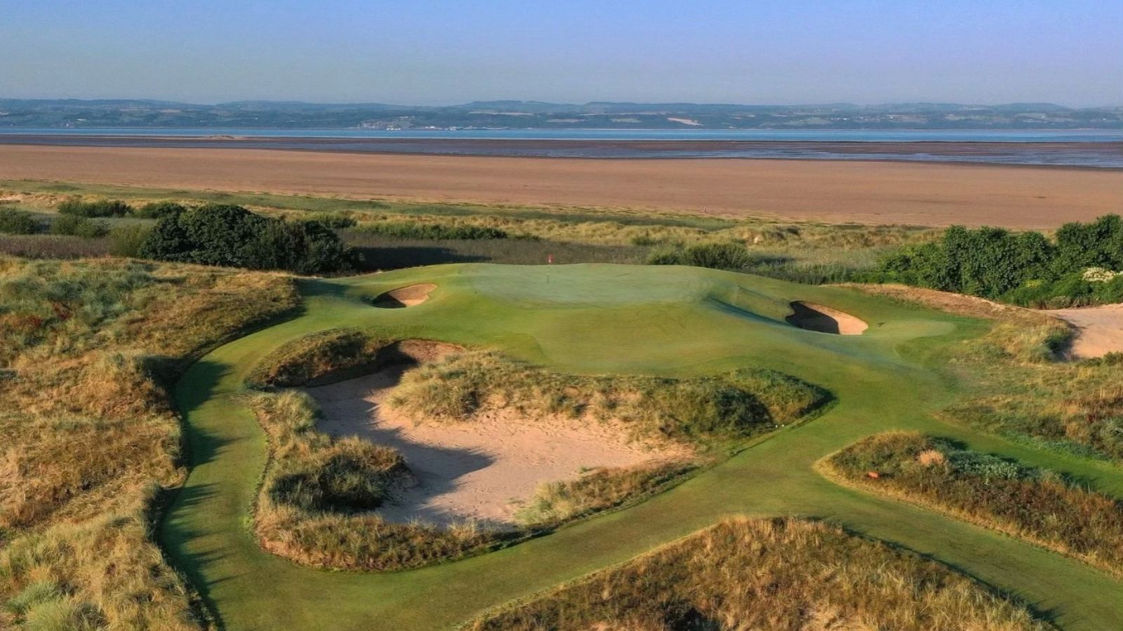 Golf course green beside sandy dunes and a lake under a clear sky