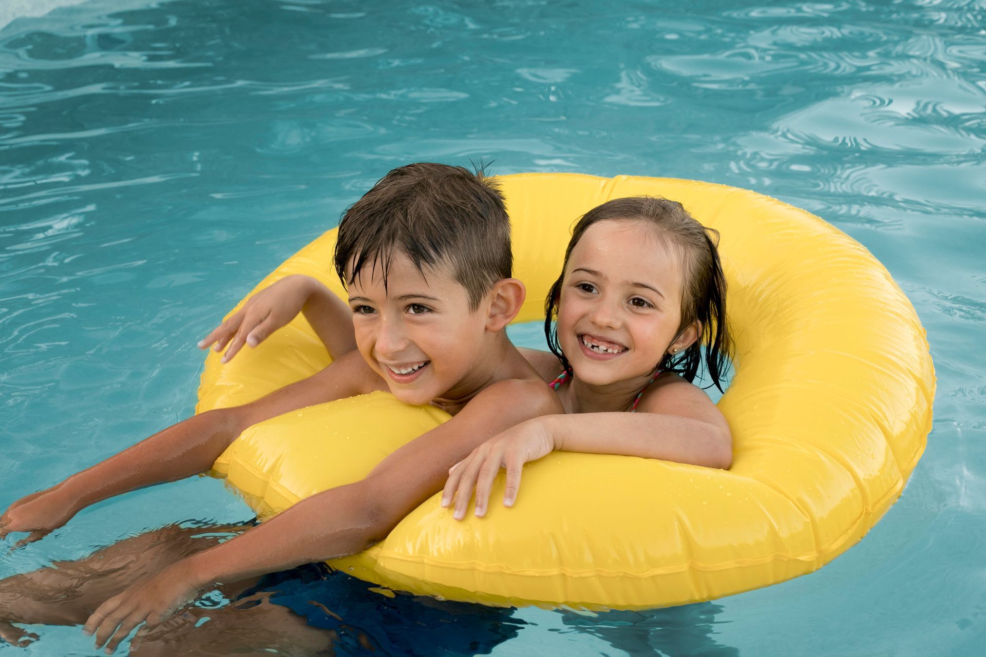 A boy and a girl are floating on a yellow life preserver in a swimming pool.