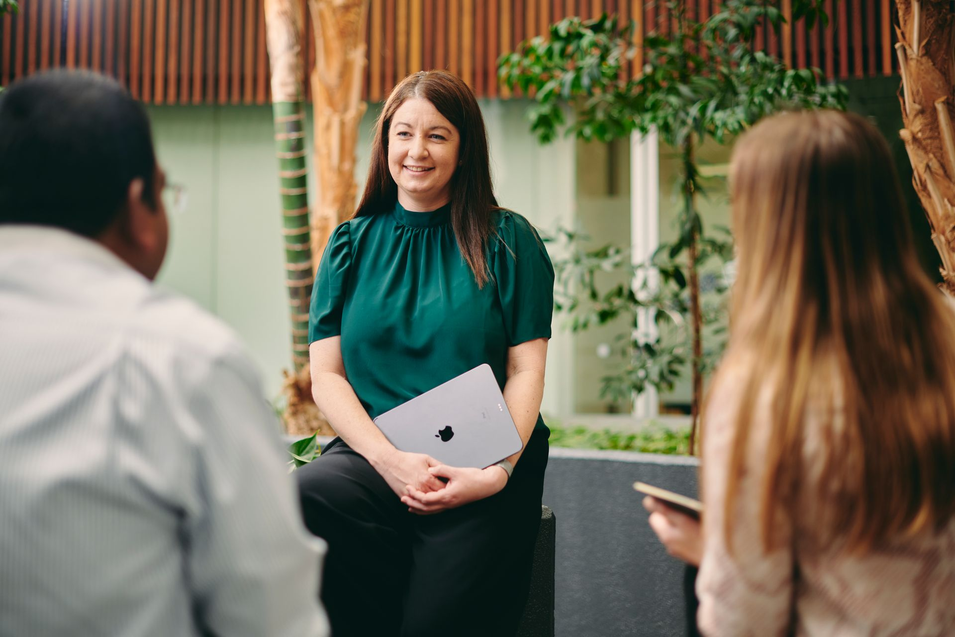 Woman holding a laptop speaks to two seated people in a modern meeting room with plants.