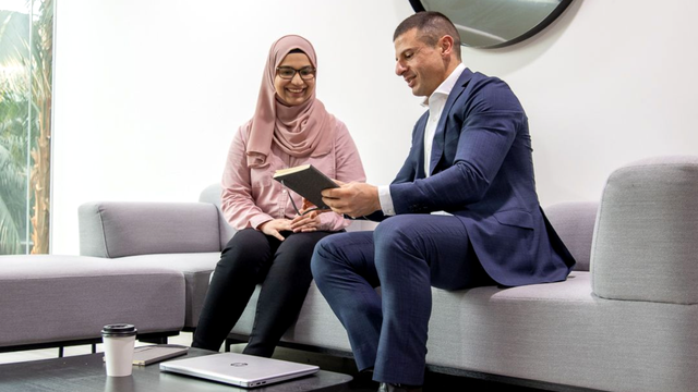 Two professionals sit on a gray sofa, reviewing a tablet in a bright office lounge.