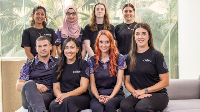 Eight people in matching black shirts posing together on a couch in a bright office lounge