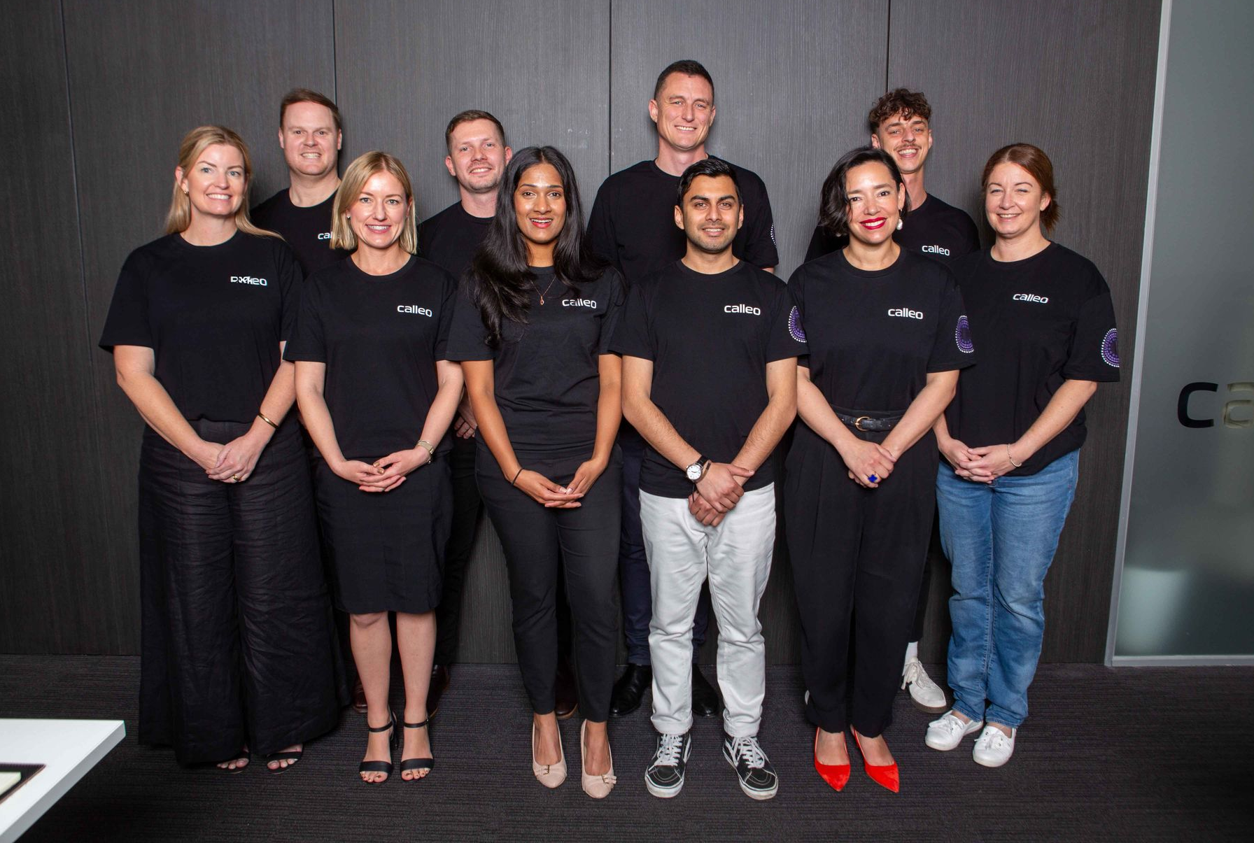 Eight people in matching black shirts posing together on a couch in a bright office lounge