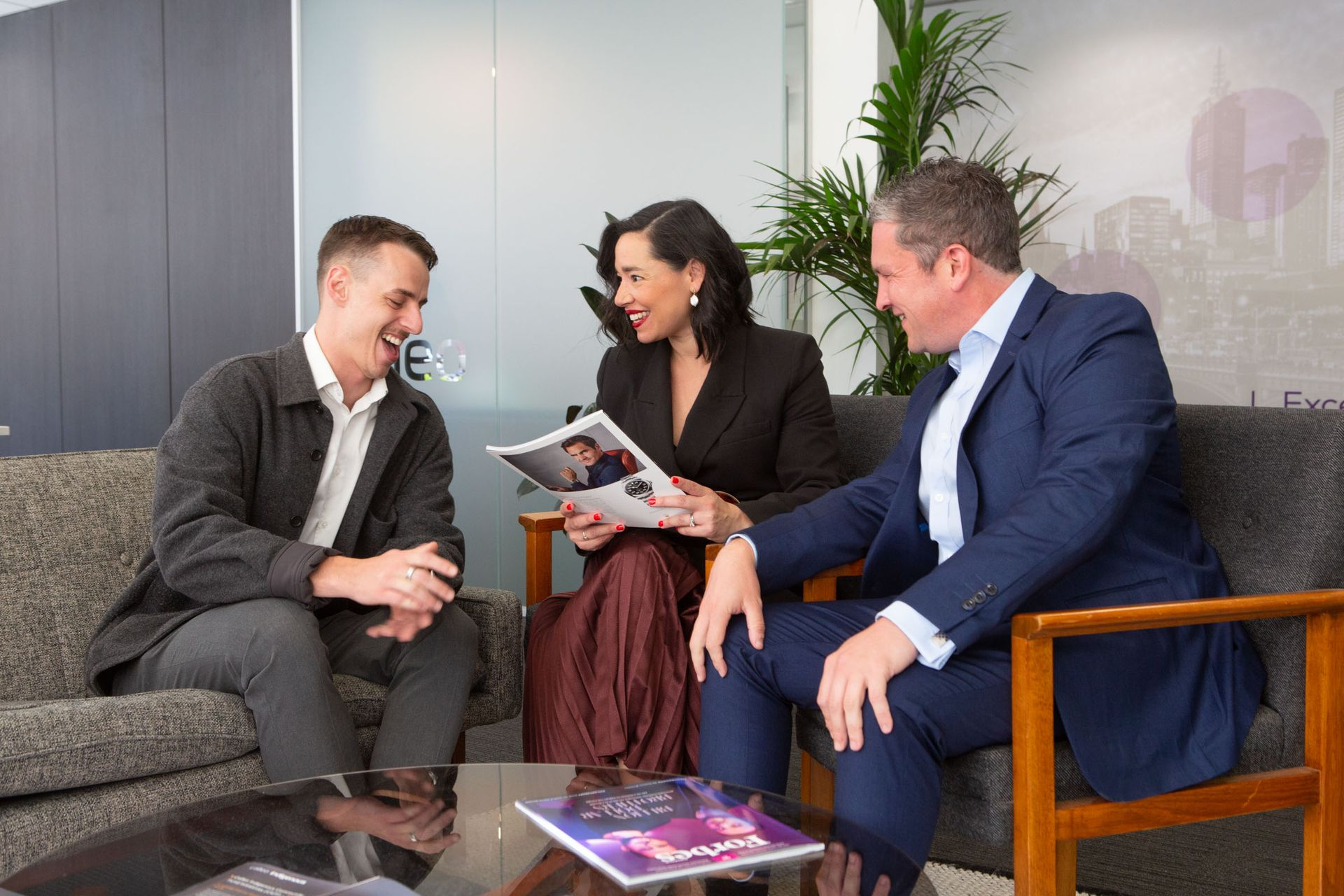 Three people in a meeting room smiling and discussing a magazine with coffee on the table