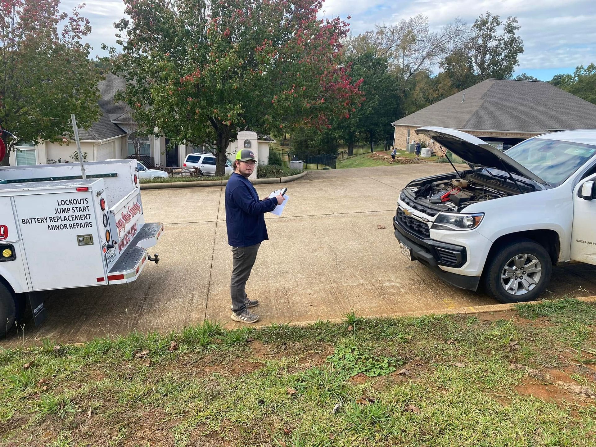 Person standing beside a white truck with hood open, holding tools in a driveway near a utility trailer.