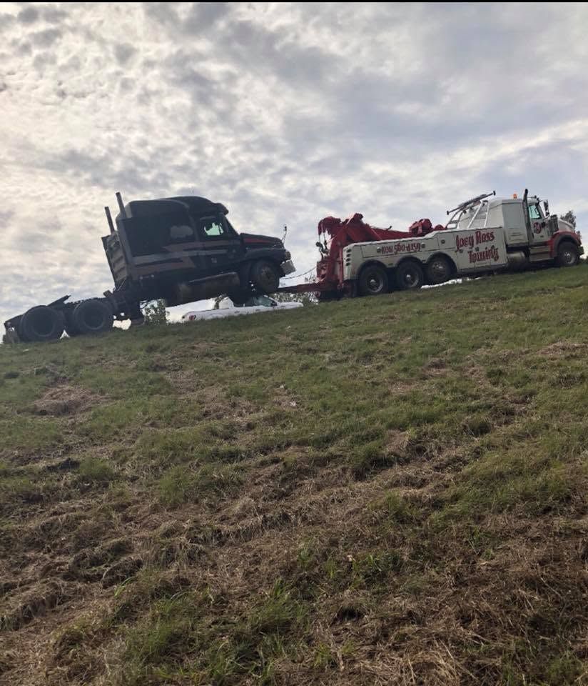Black tow truck hauling a damaged truck on a grassy roadside under cloudy sky