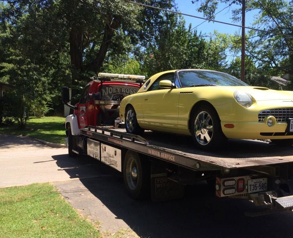 Yellow sports car on a flatbed tow truck parked beside a tree-lined street.