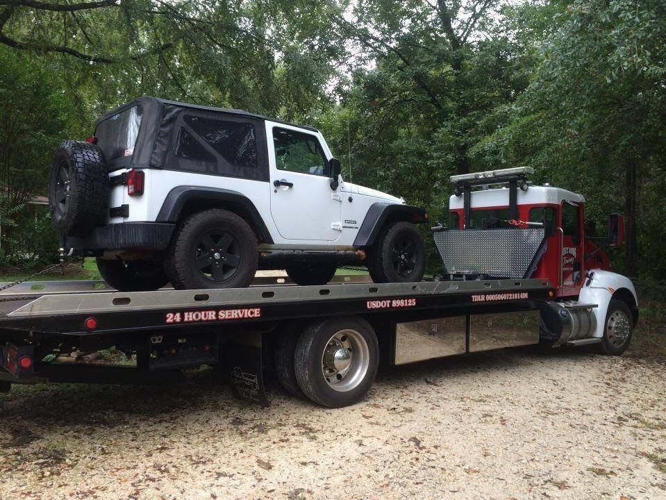 Tow truck carrying a white Jeep Wrangler and a red buggy on a gravel road in a wooded area