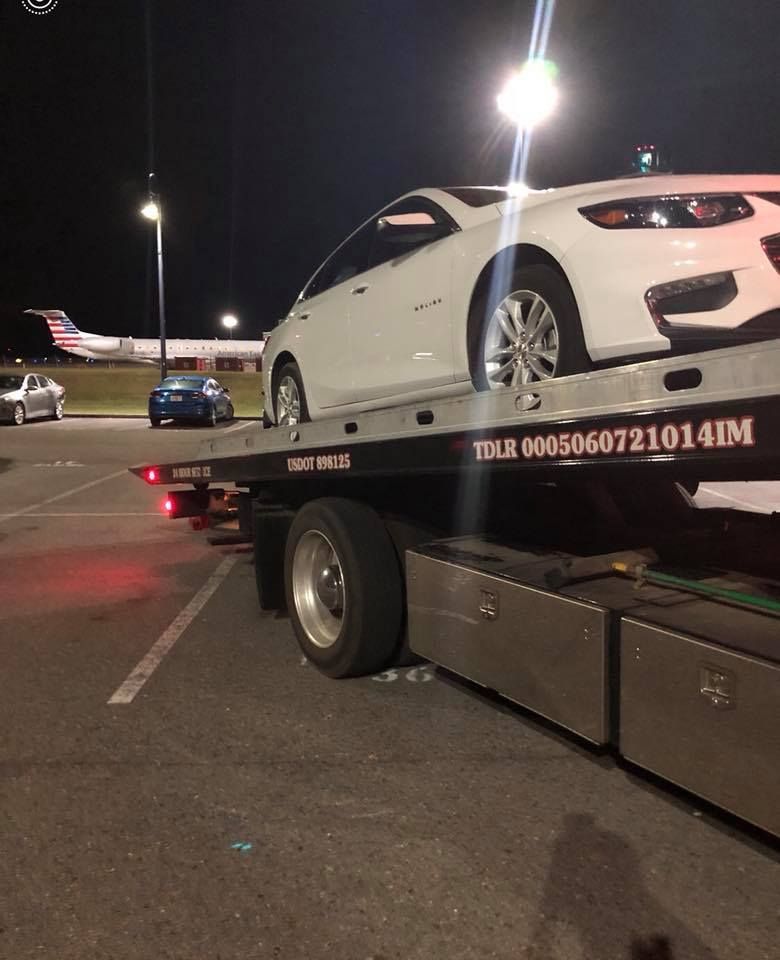 White sports car on a tow truck at night on a road, with streetlights and other cars nearby