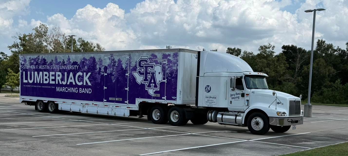 White semi-truck with a purple “Lumberjack” trailer parked in a lot under a cloudy sky