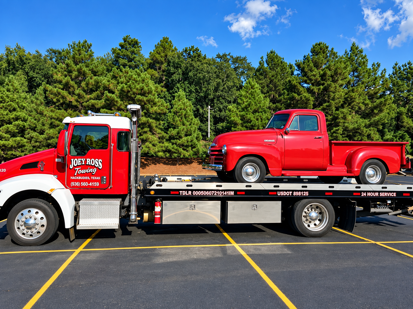 Red pickup truck secured on a flatbed tow truck in a parking lot, with trees and blue sky behind.