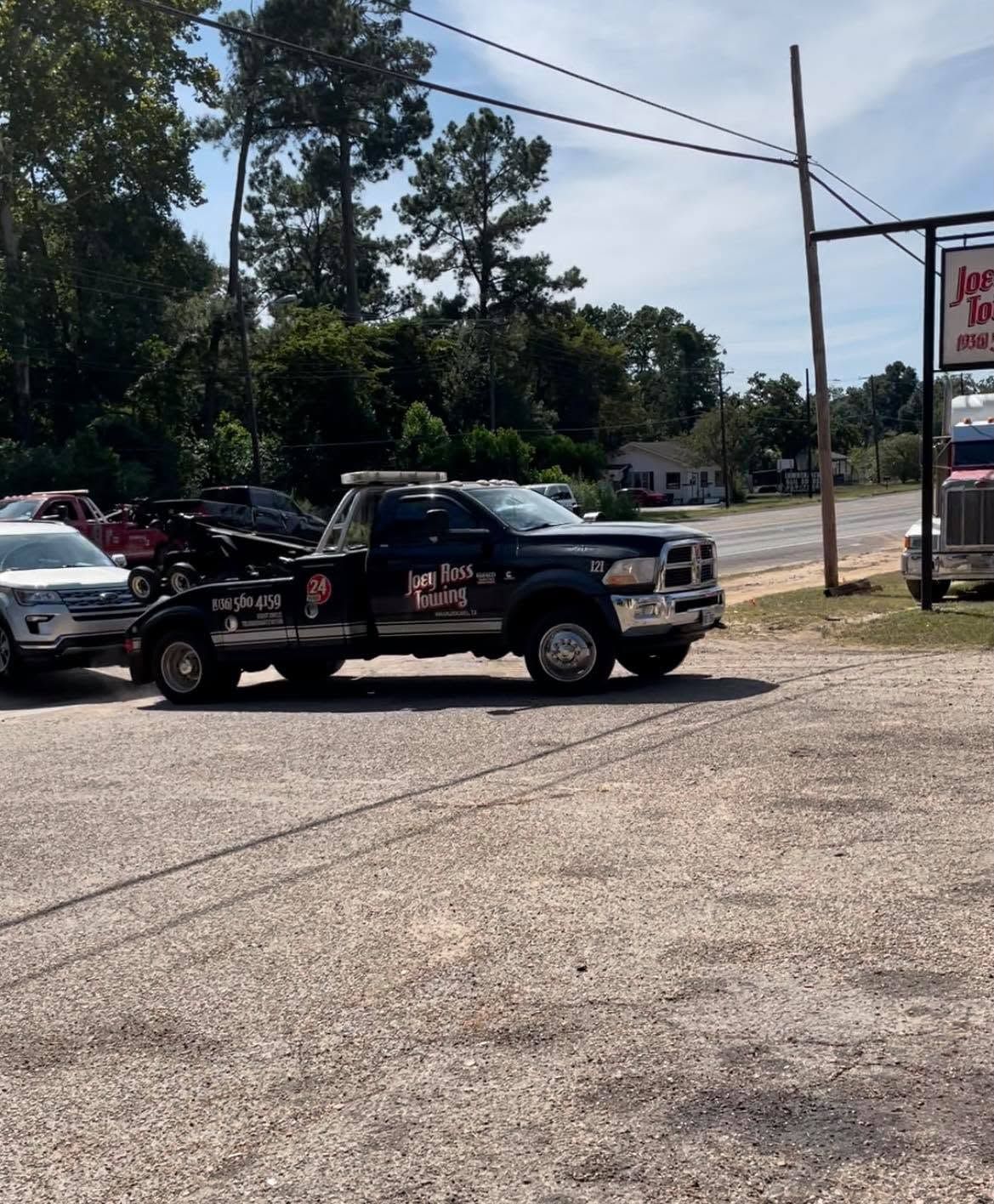 Tow truck with a black pickup hauling a car on a gravel lot under a sunny sky