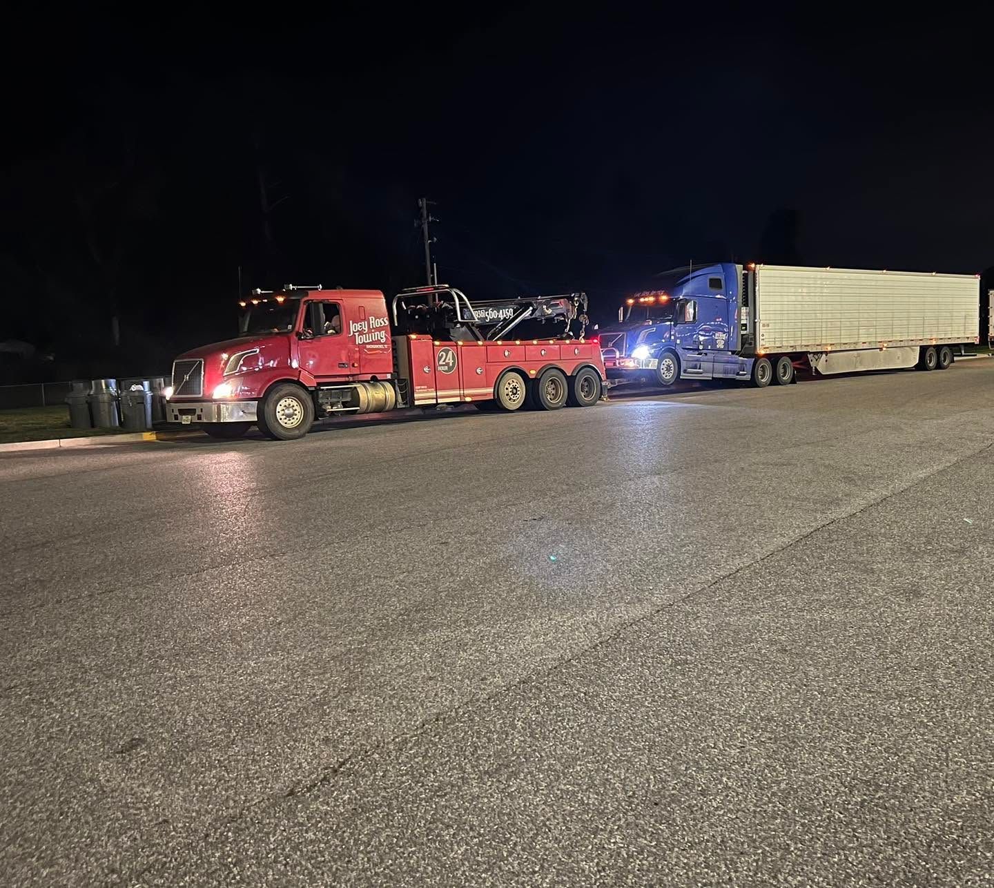 Red tow truck towing a blue semi-truck and trailer at night on a road