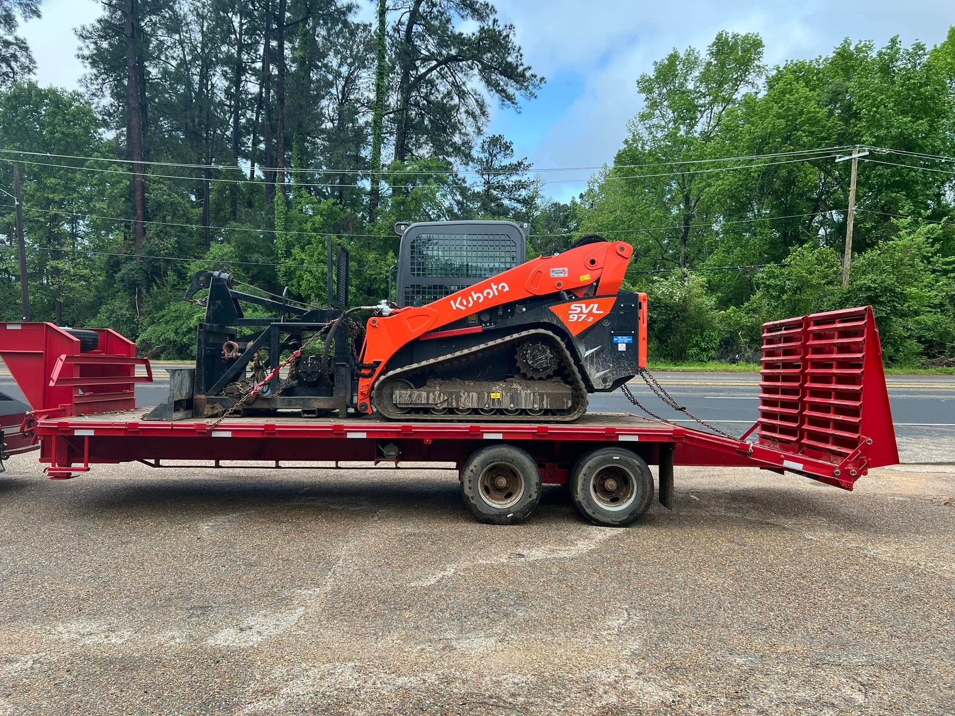 Orange compact track loader on a red trailer in a gravel lot with trees in the background