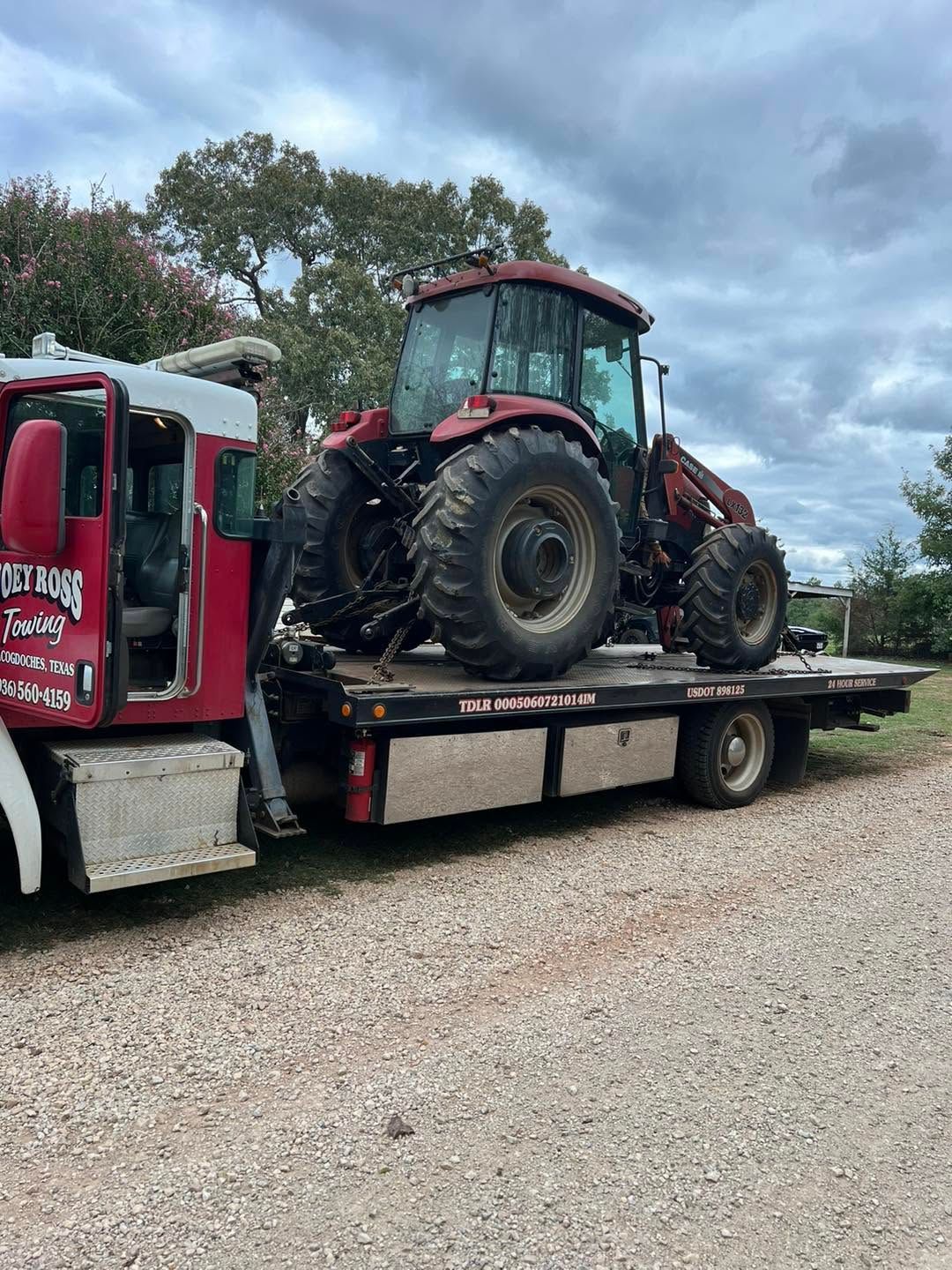 Red flatbed tow truck hauling a red tractor on a gravel lot under cloudy skies