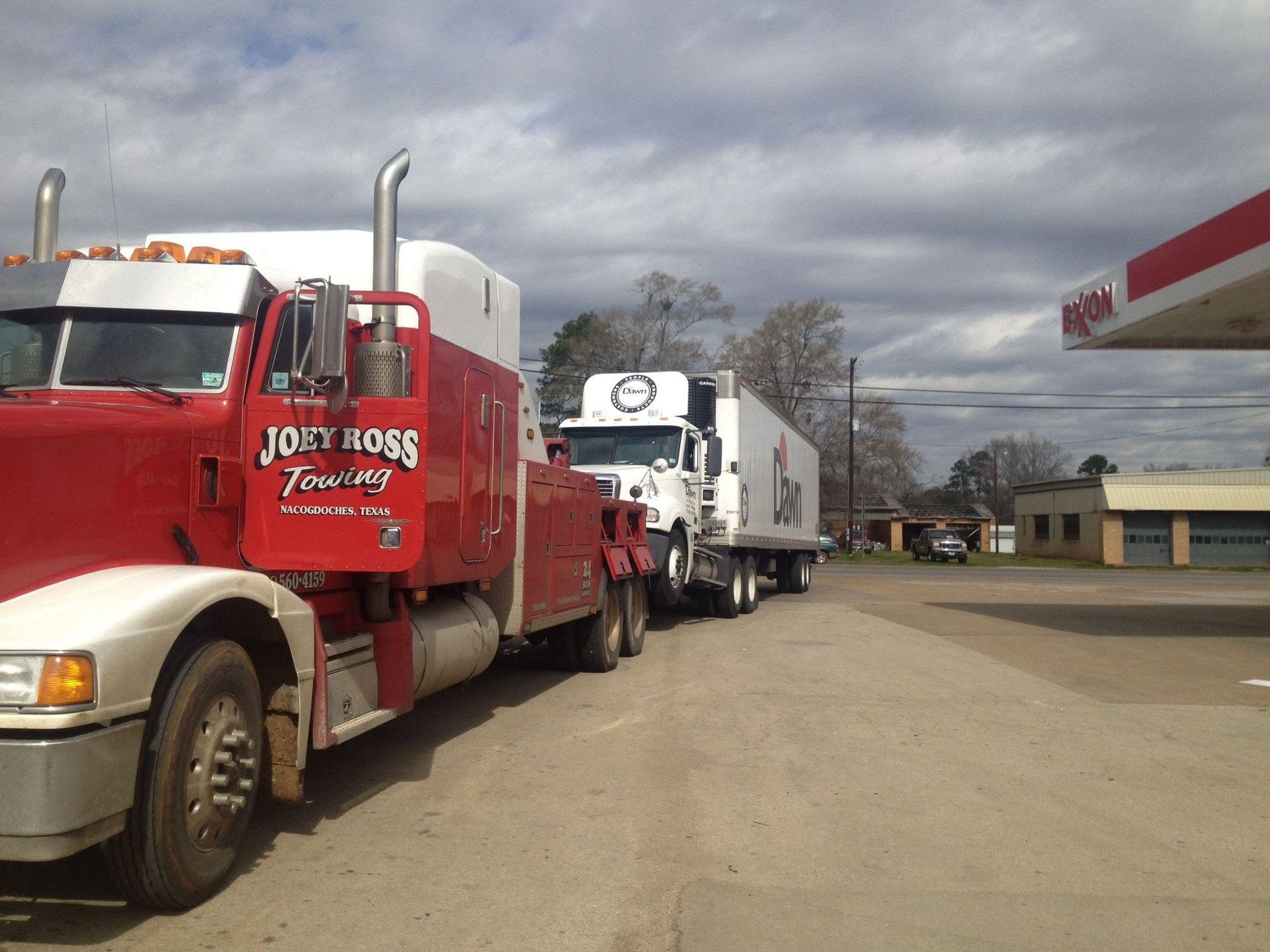 Two parked semi trucks, red and white, lined up beside a gas station under a cloudy sky.