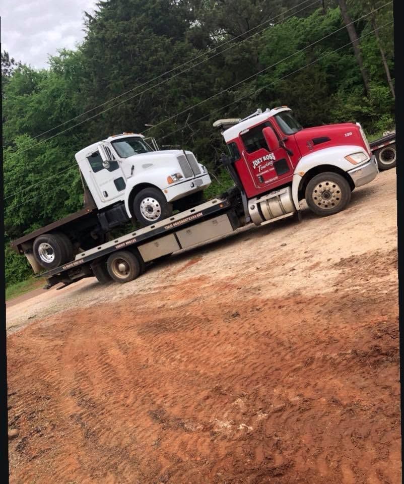 Red tow truck hauling a white flatbed truck on a dirt road beside trees.