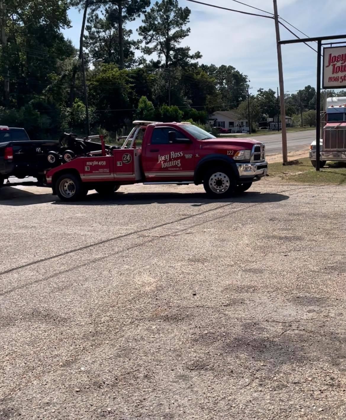 Red tow truck parked on a gravel lot near a road, with trees and a roadside sign in the background.